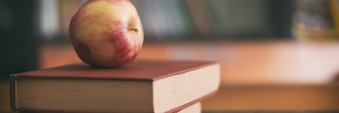 Apple sitting on a pile of books on a classroom desk.