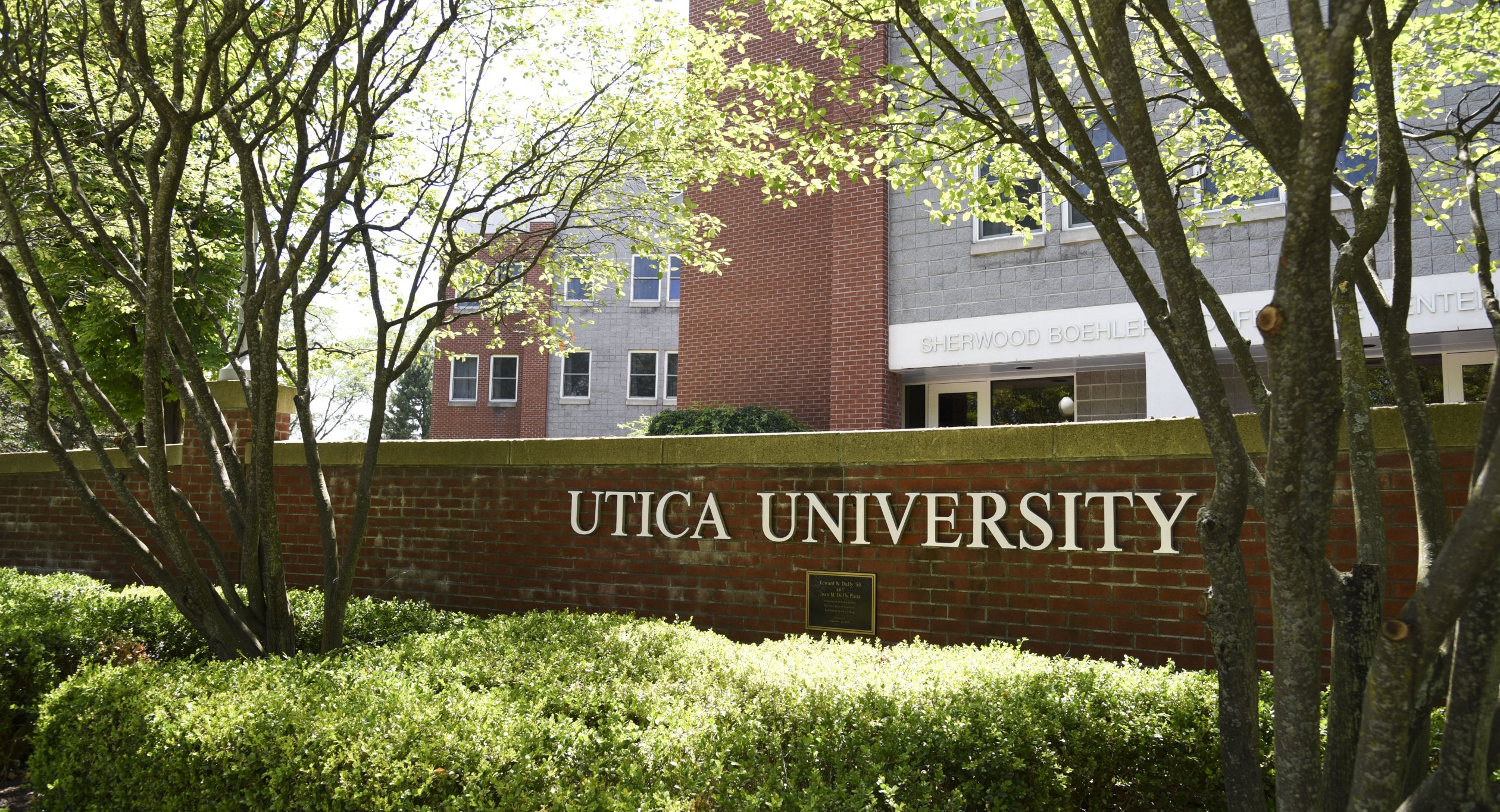 The words Utica University against a brick wall in front of a residence hall.