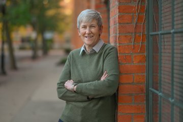 Stephanie Nesbitt in a green sweater, leans against a brick wall with her arms folded.