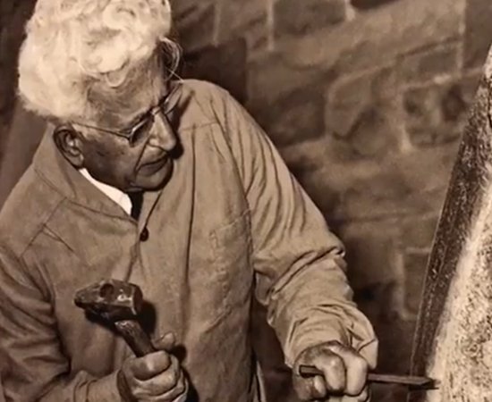 Black and white image of Henry Dispirito at work on a sculpture, with hammer and chisel in hand.