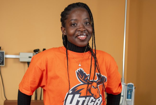 Lois Mensah, in an orange Utica t-shirt, stands against an examination table in the nursing lab.