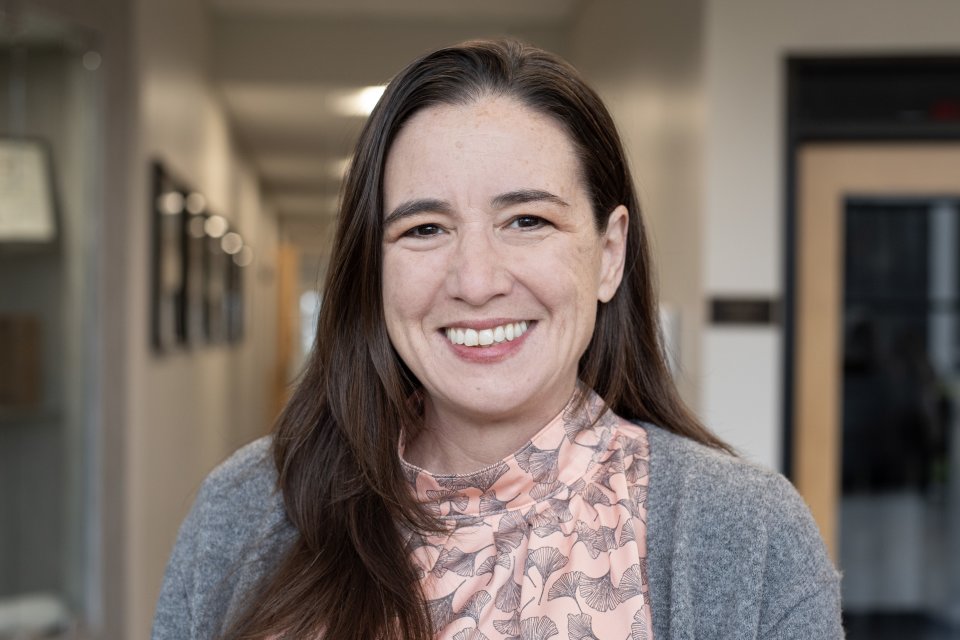 Deborah Pollack, with long brown hair, pink top and gray sweater, smiles at camera.